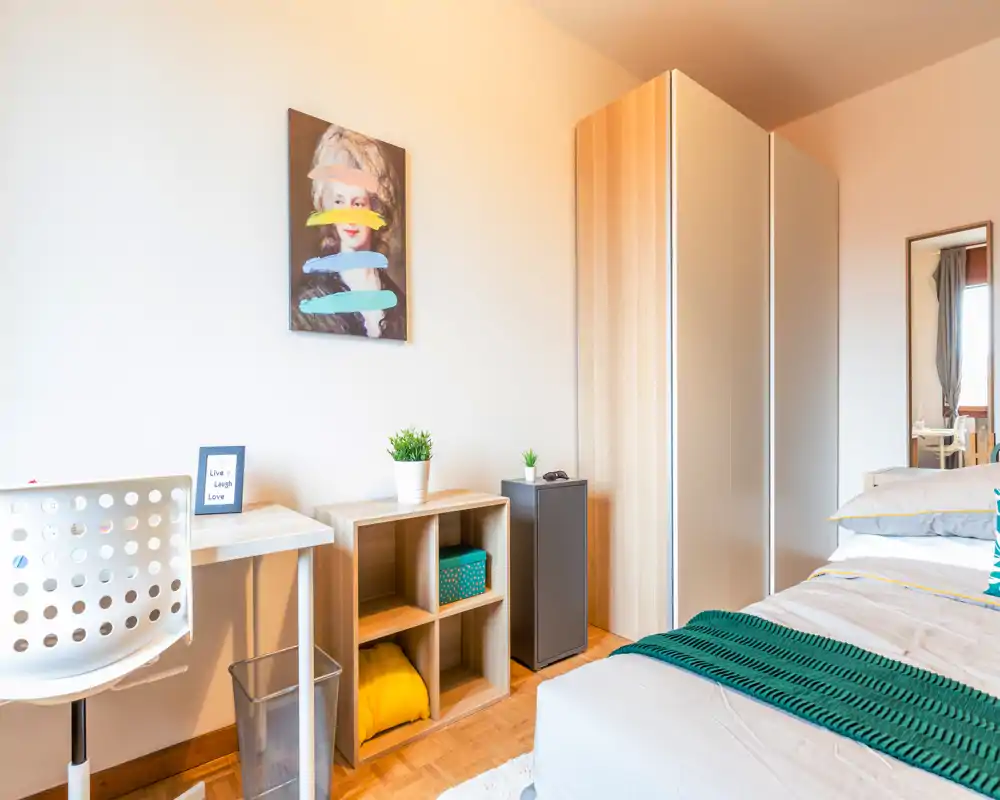 Bright, modern bedroom with a neatly made bed, wooden wardrobe and a small desk area — ideal for a comfortable stay with natural light and cozy textiles.