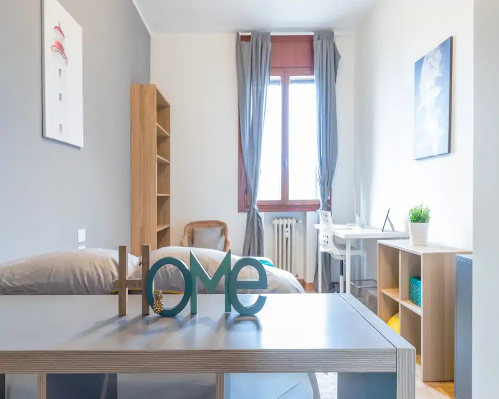 Bright, neatly styled bedroom with a comfortable double bed, desk by the window and tasteful wooden shelving — ideal for a cozy stay or a home office setup.