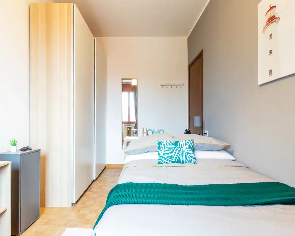 Bright, neatly staged bedroom with a double bed, decorative cushions and a teal throw. The room features a large wardrobe, bedside table and minimalist décor, ideal for rental listings highlighting cleanliness and comfort.