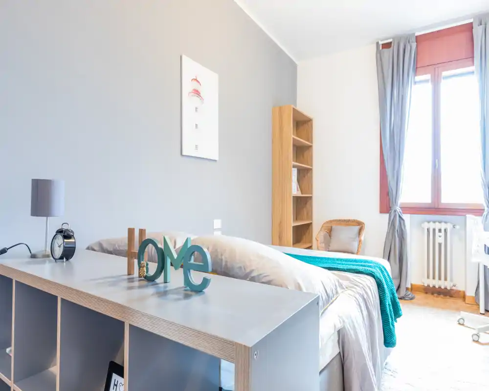 Bright, modern bedroom with a neatly made double bed, turquoise throw and simple shelving; a large window fills the room with natural light.
