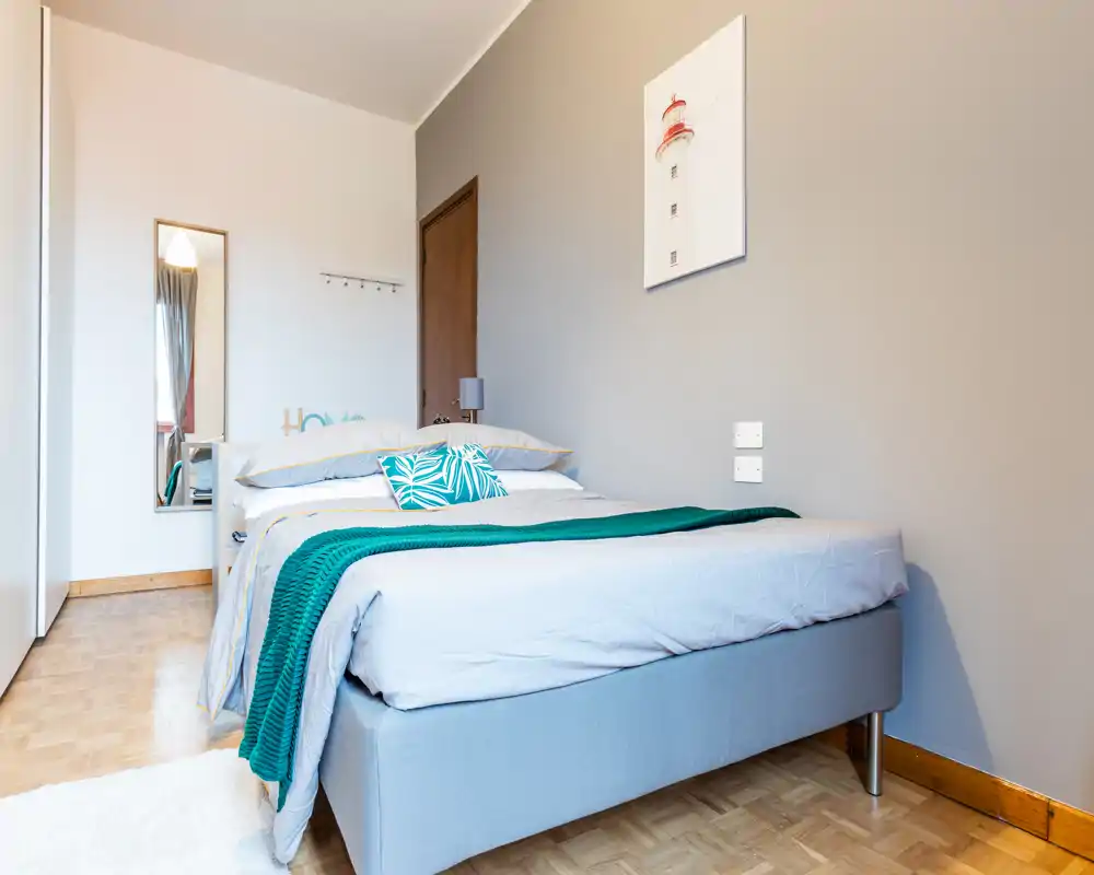 Bright, modern bedroom with a neatly made double bed, teal throw and decorative cushions, soft wooden floor and minimalist wall art — ideal for rental listings highlighting comfort and style.