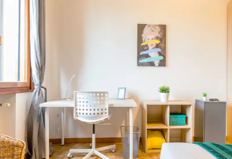 Bright, simply styled bedroom corner with a white desk, modern perforated chair and a neatly made bed with colorful throw; natural light from a window and minimalist shelving create an airy, welcoming feel.