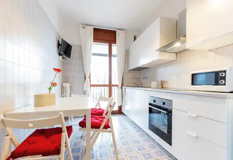 Bright, modern kitchen with white cabinets, built-in oven and a dining table with red-cushioned chairs next to a balcony door letting in natural light.
