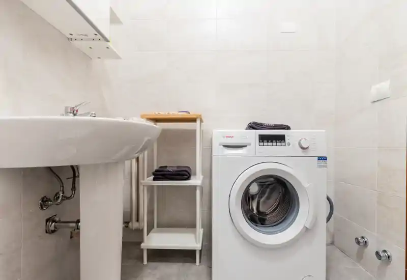 Compact bathroom-laundry area with a front-loading washing machine, pedestal sink and small shelving; bright, tiled walls and clean contemporary finishes.