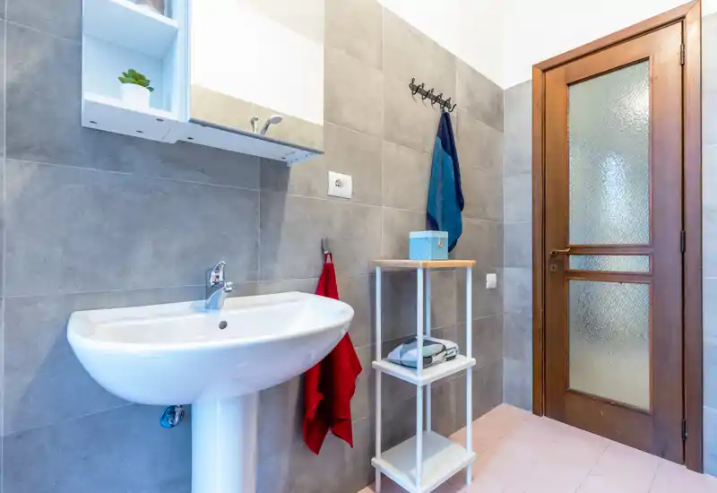 Bright, modern bathroom with pedestal sink, wall-mounted mirror cabinet, towel hooks and a wooden door — clean and functional space.