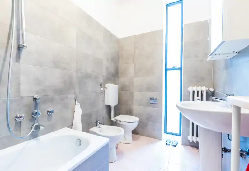 Bright, modern bathroom with a bathtub, bidet, toilet and pedestal sink; light gray wall tiles and a tall narrow window provide natural light.