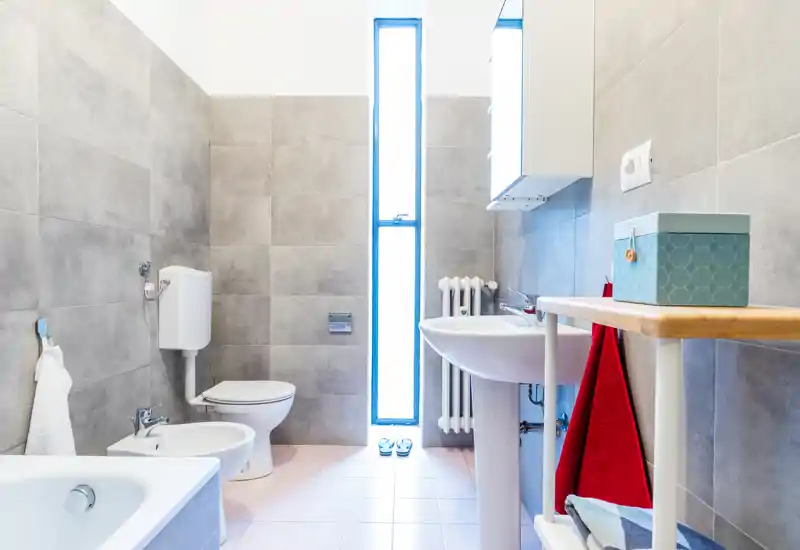 Bright, modern bathroom with toilet, bidet, sink and bathtub. Neutral grey tiles and a tall narrow frosted window provide natural light and a clean minimalist look.