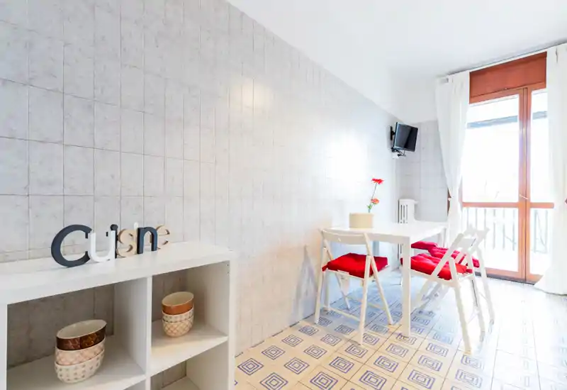 Bright, compact kitchen/dining area with a small table, chairs with red cushions and a balcony door letting in abundant natural light — clean, modern styling ideal for a small apartment.
