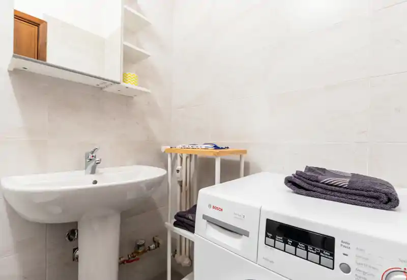 Compact bathroom with sink, mirrored cabinet and a washing machine; light tiled walls and neat storage, ideal for functional stays.