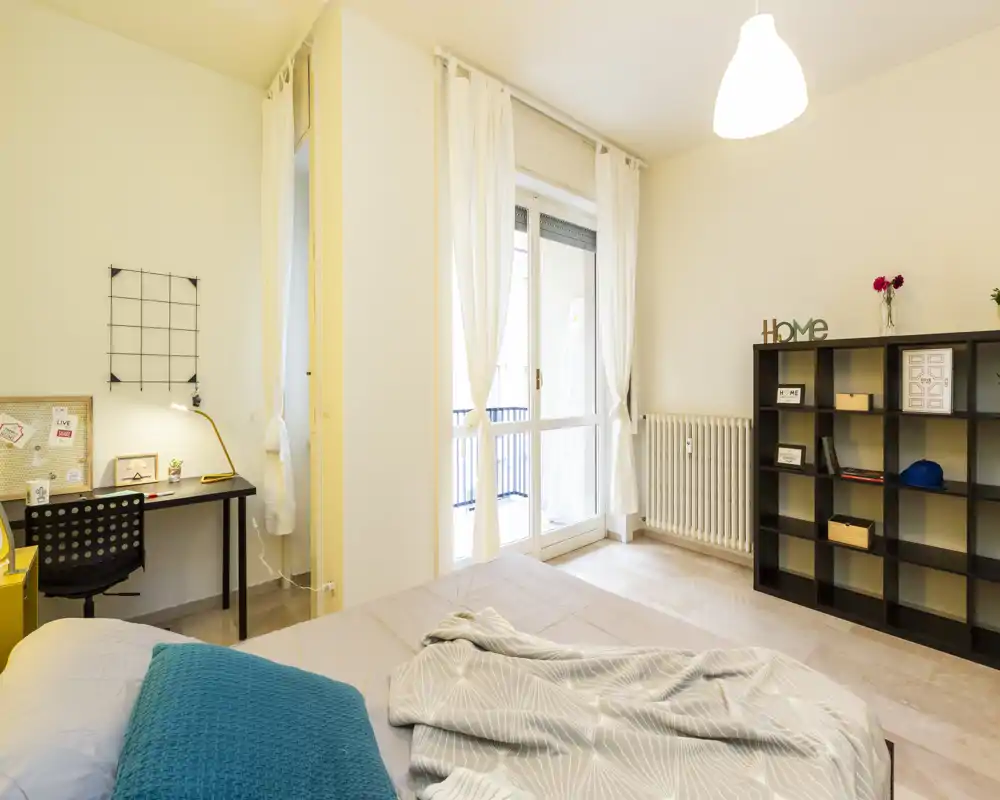 Sunlit bedroom with a neatly made double bed, soft textiles and a work desk by the window. Bright, modern styling with shelving for storage and a balcony door providing natural light.
