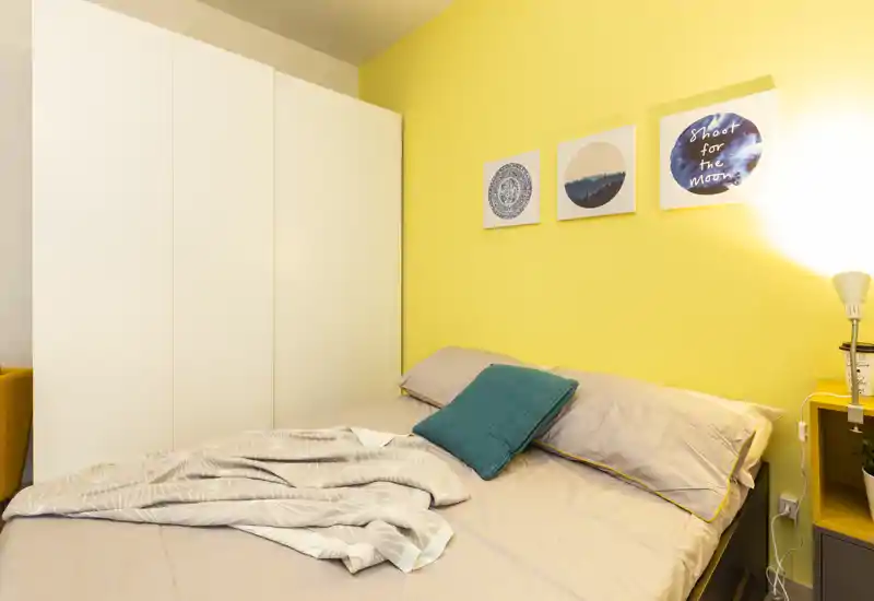 Bright, cozy bedroom with a large bed, teal cushion and warm yellow accent wall; white wardrobe and bedside lamp complete the scene.