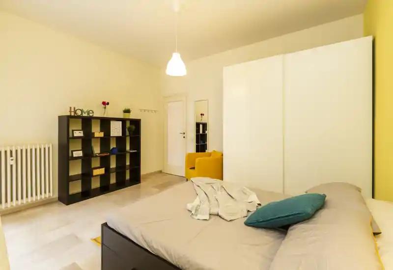 Bright, modern bedroom with a comfortable double bed in the foreground, a large white wardrobe and a cozy yellow armchair. The room features clean lines, neutral tones and decorative accents for a welcoming atmosphere.