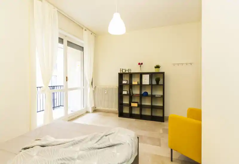 Bright, simply styled bedroom with a large sliding balcony door, a low bed with soft linens, shelving unit and a yellow accent armchair—ideal for a cozy one-bedroom rental.