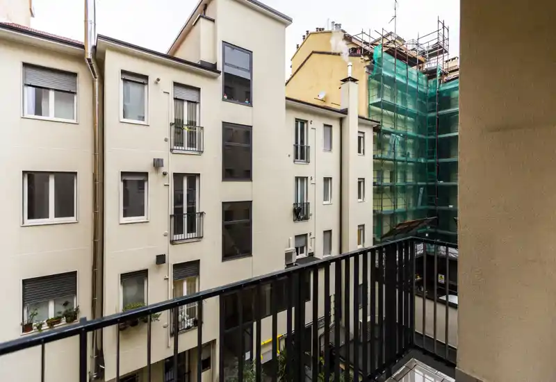 Small private balcony overlooking neighboring apartment facades and a courtyard; metal railing, city view with nearby scaffolding and muted daylight.