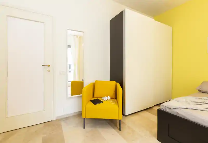 Bright, modern bedroom corner with a yellow accent armchair and a partially visible bed. Clean lines, a large wardrobe and warm tones create an inviting, contemporary sleeping space.