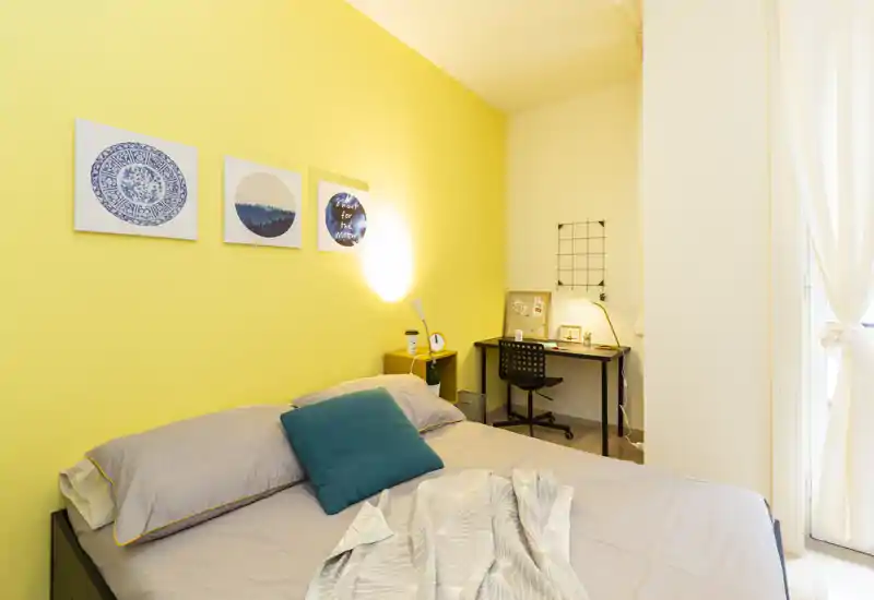 Chambre lumineuse et chaleureuse avec un grand lit contre un mur jaune, lampe de chevet et un petit bureau dans le coin — idéale pour se reposer et travailler légèrement.