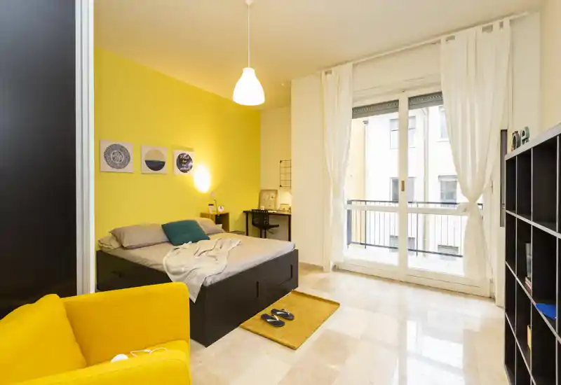 Sunny, modern bedroom with a comfortable double bed, bright yellow accent wall, workspace and balcony access. Clean lines and warm textiles create an inviting atmosphere ideal for rental photos.