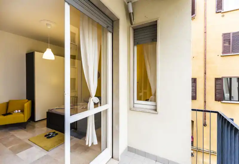 Sunny bedroom seen from the balcony: a neatly made bed with light curtains, a yellow armchair and warm pendant lighting create a cozy city-room atmosphere.