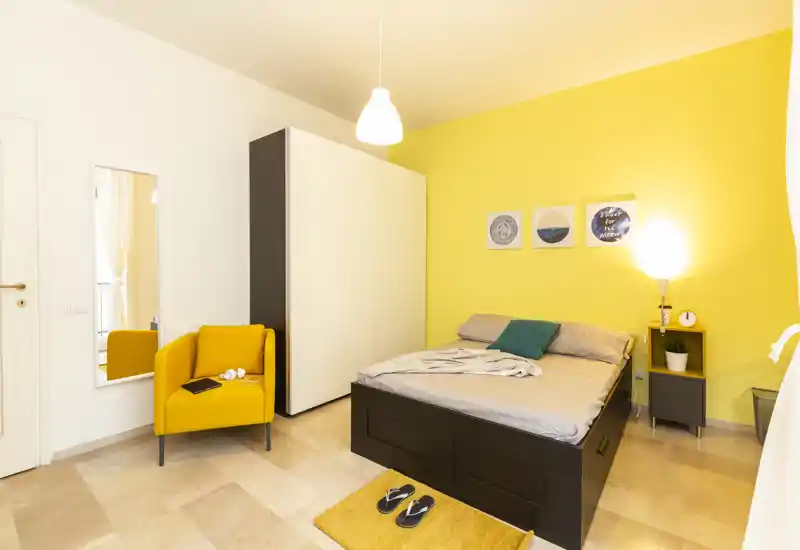 Bright, modern bedroom with a double bed, bedside table and a cozy armchair against a cheerful yellow accent wall — ideal for a stylish short-term rental.