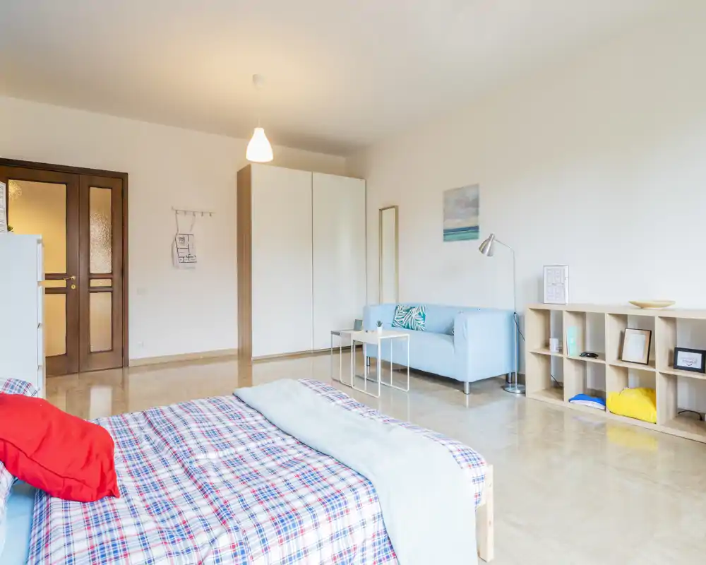 Camera da letto luminosa e moderna con un letto in primo piano coperto da una coperta a quadri e un divano azzurro sullo sfondo; linee pulite, arredo minimale e luce naturale rendono lo spazio invitante per gli annunci.