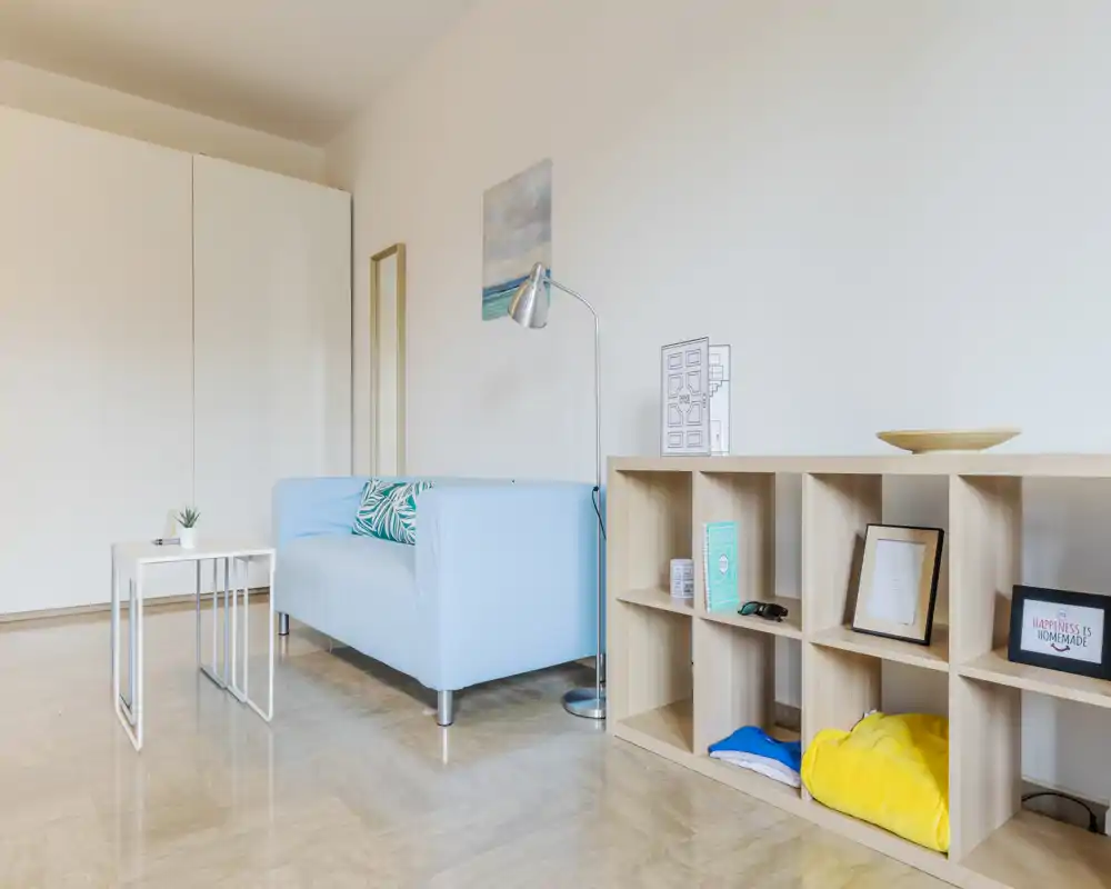Zona living luminosa e minimale con divano azzurro chiaro, scaffalatura moderna e tavolini semplici — linee pulite e accenti morbidi creano un ambiente rilassante.