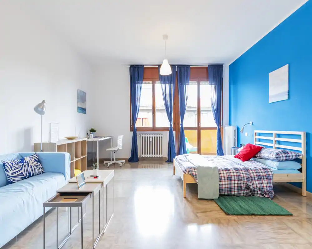 Camera luminosa e moderna in stile monolocale con un comodo letto singolo contro una parete blu vivace, area soggiorno accogliente e scrivania vicino alle grandi finestre che illuminano l'ambiente.