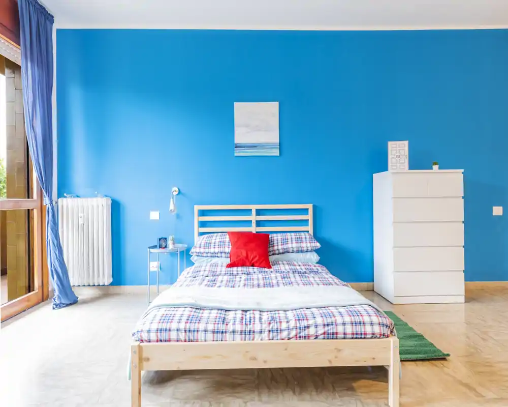 Camera da letto luminosa e moderna con letto matrimoniale in legno, biancheria a quadri e una parete principale blu intensa. Ampia finestra che garantisce molta luce naturale e uno stile minimale adatto agli annunci.
