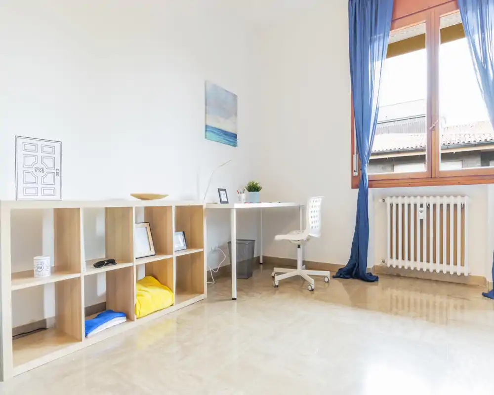 Angolo di camera luminoso e minimale adibito a zona lavoro, con scrivania semplice, scaffale a cubi e una grande finestra che illumina l'ambiente.