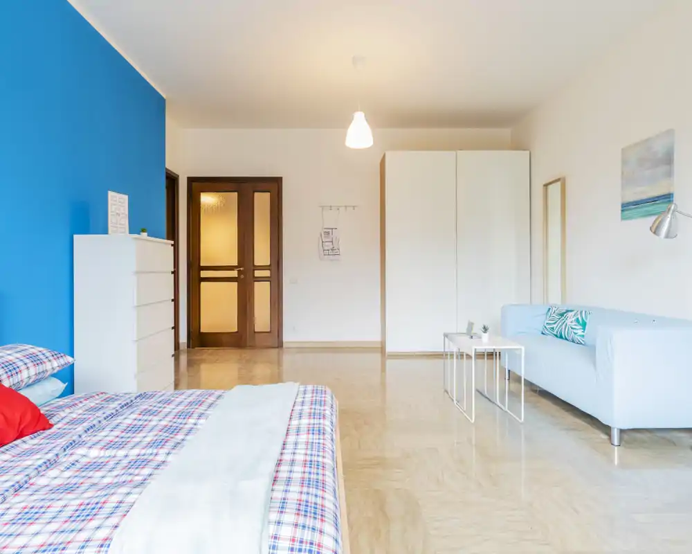 Ampia e luminosa camera da letto con un grande letto in primo piano, parete accentata blu e una piccola zona salotto. Arredo moderno e pulito con molta luce, perfetta per annunci di affitto che vogliono valorizzare comfort e stile.
