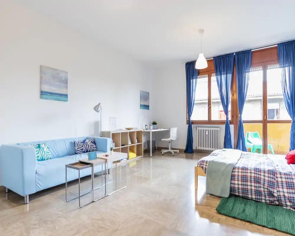 Camera da letto luminosa e ben arredata con letto matrimoniale, divano e angolo studio; ampie finestre con tende blu garantiscono molta luce naturale e un’atmosfera accogliente e ariosa.