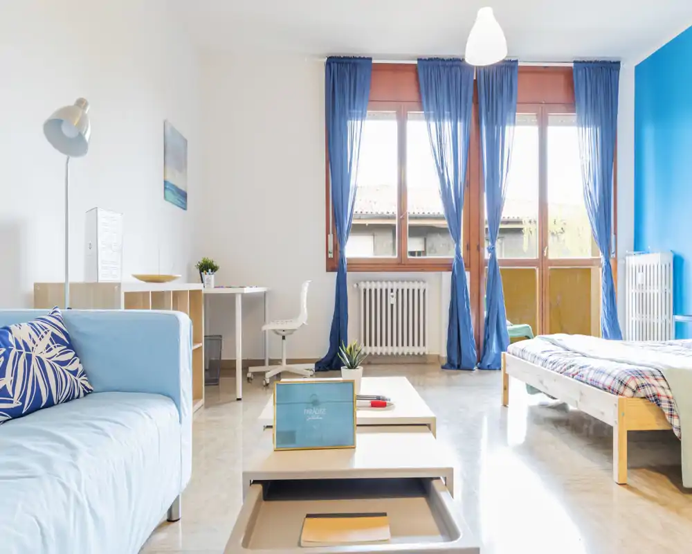 Camera luminosa e accogliente con letto matrimoniale, divano e ampia finestra che lascia entrare molta luce naturale. Arredamento moderno e minimale con accenti blu e una postazione di lavoro ordinata, perfetta per affitti brevi.