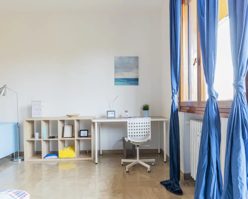 Area studio/soggiorno luminosa e minimal con scrivania bianca, sedia da ufficio forata e un piccolo mobile a cubi. Ampia finestra con tende blu illumina la stanza; arredamento semplice con una pianta e un quadro marino.