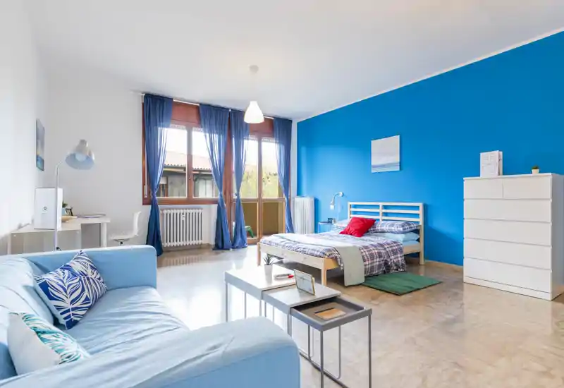 Ampia e luminosa camera da letto con parete accentuata blu, letto matrimoniale, divano comodo e grandi finestre che lasciano entrare molta luce naturale — perfetta per un annuncio accogliente.