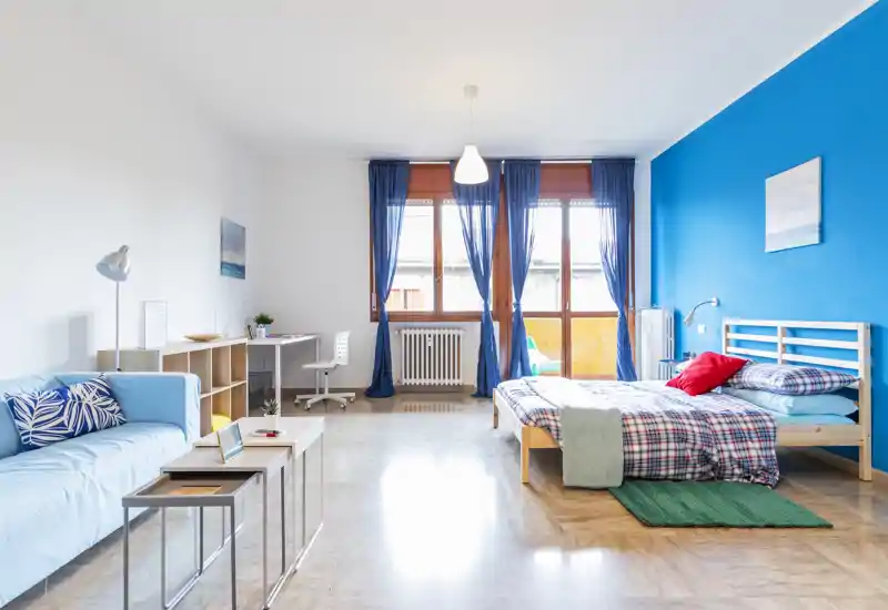 Camera luminosa e moderna in stile monolocale con un comodo letto singolo contro una parete blu vivace, area soggiorno accogliente e scrivania vicino alle grandi finestre che illuminano l'ambiente.