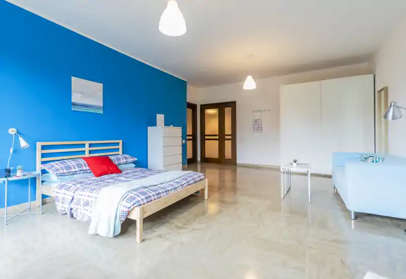 Ampia e luminosa camera da letto con letto matrimoniale in legno su una parete blu a contrasto, biancheria con fantasia e una zona soggiorno. La stanza è ben illuminata, arredata con cura e ideale per annunci che puntano su spazio e stile moderno.