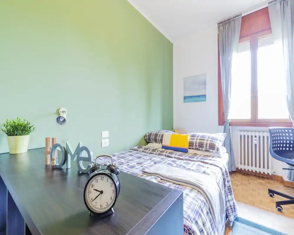 Camera da letto luminosa e ordinata con letto matrimoniale in coordinato scozzese, cuscini e una grande finestra che illumina la stanza. Un piano scrivania con oggetti decorativi e una sveglia aggiunge funzionalità.