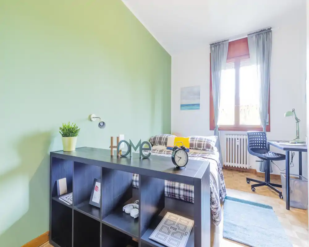 Camera da letto luminosa e ben arredata con letto matrimoniale, biancheria a quadri e scrivania vicino alla finestra; parete verde a contrasto e mobile contenitore per ottimizzare lo spazio.
