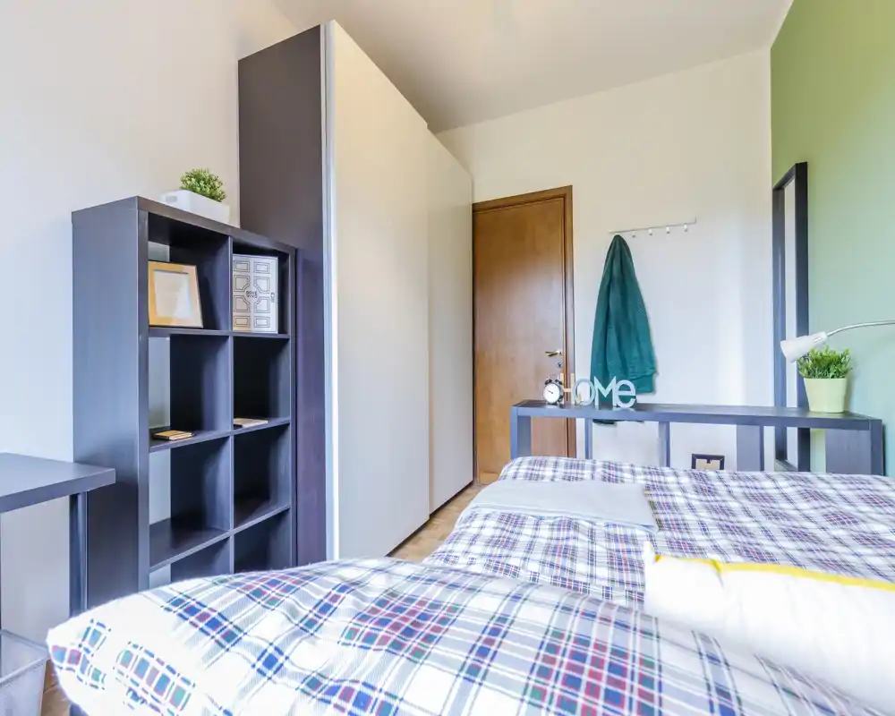 Camera da letto luminosa e ben allestita con letto matrimoniale a fantasia in primo piano, grande armadio e scaffalatura per riporre, con semplici dettagli decorativi che la rendono accogliente.