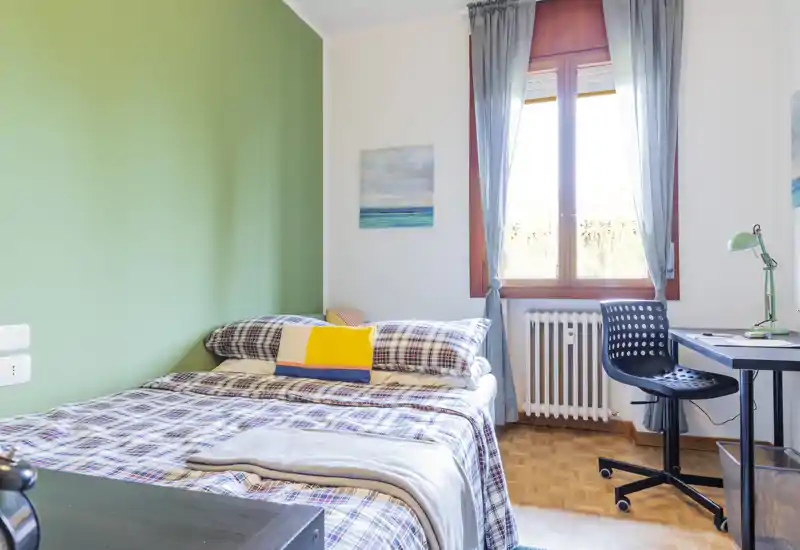 Camera da letto luminosa e accogliente con letto matrimoniale in lino a quadri, cuscino colorato e una scrivania vicino alla finestra che illumina l’ambiente con luce naturale.