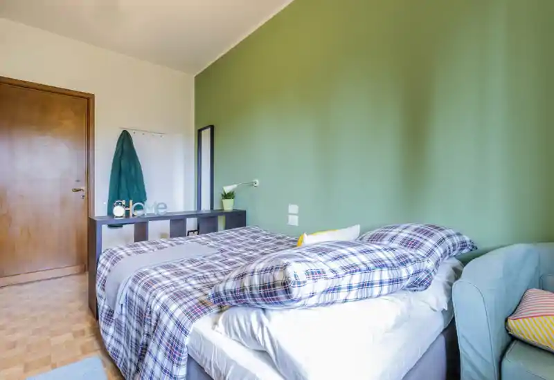 Camera da letto luminosa e ordinata con letto matrimoniale in piumoni a quadri contro una parete verde; la luce naturale e i tessuti morbidi creano un ambiente accogliente.