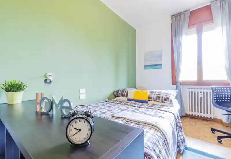 Camera da letto luminosa e ordinata con letto matrimoniale in coordinato scozzese, cuscini e una grande finestra che illumina la stanza. Un piano scrivania con oggetti decorativi e una sveglia aggiunge funzionalità.