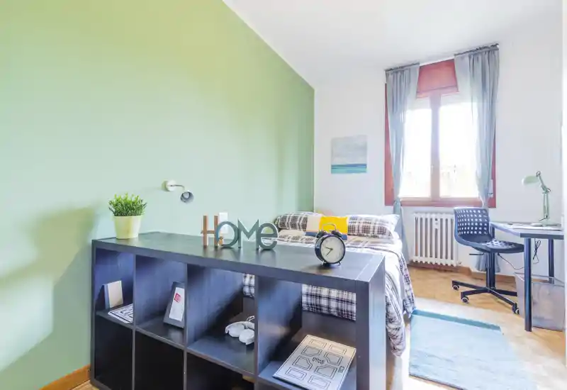 Camera da letto luminosa e ben arredata con letto matrimoniale, biancheria a quadri e scrivania vicino alla finestra; parete verde a contrasto e mobile contenitore per ottimizzare lo spazio.