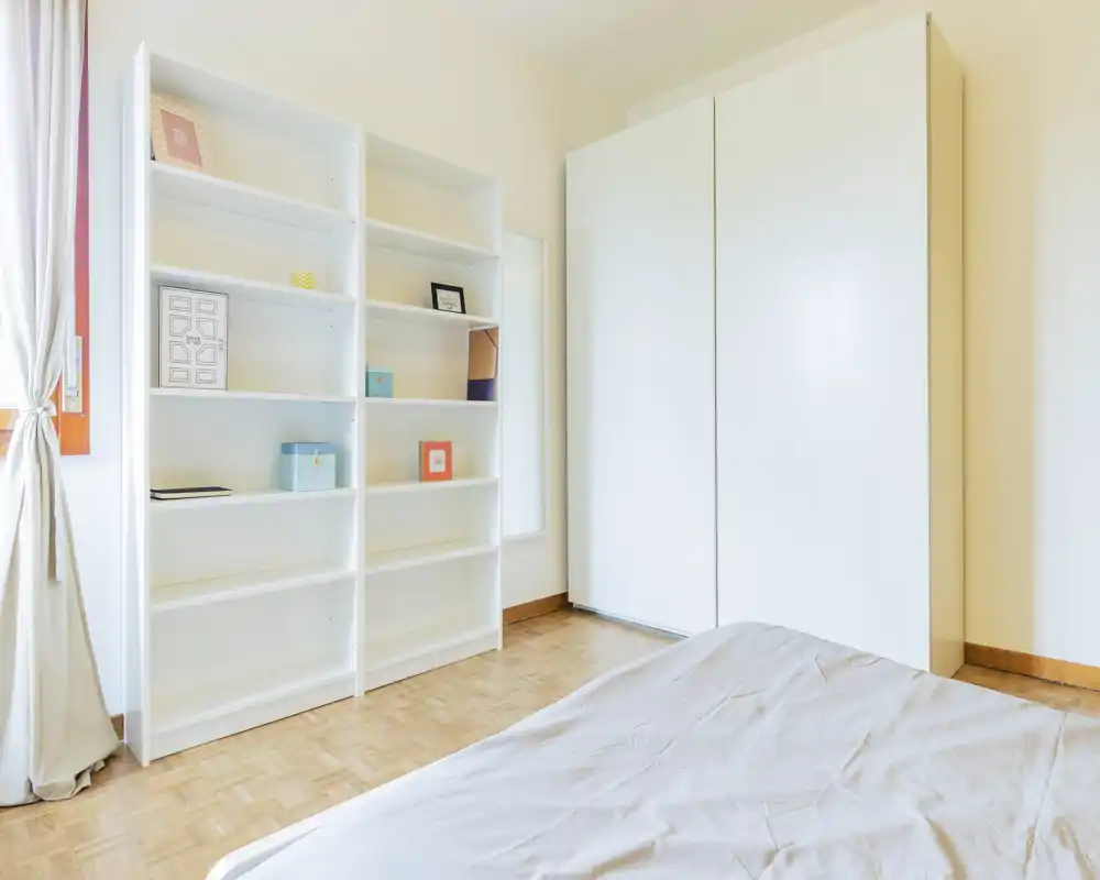 Camera da letto luminosa e minimale con il letto parzialmente visibile in primo piano, scaffali bianchi e un grande armadio bianco; parquet pulito e luce naturale dalla finestra con tenda.
