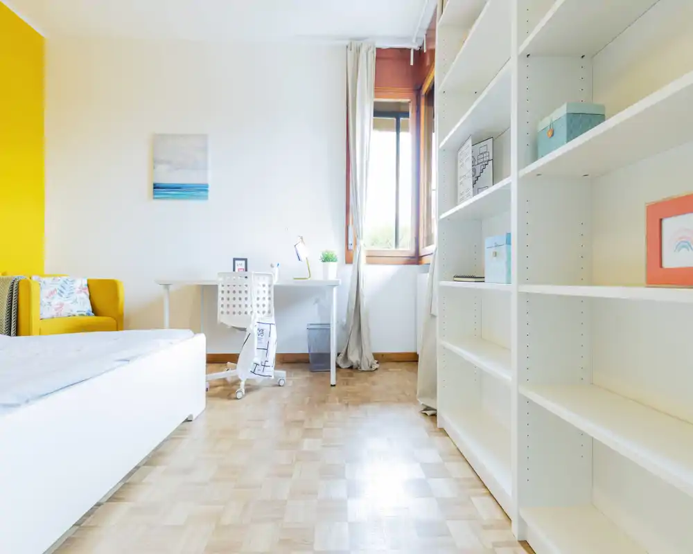 Camera da letto luminosa e ariosa con letto bianco, scrivania vicino alla finestra e libreria a parete; una poltrona gialla vivace dona un tocco di calore.