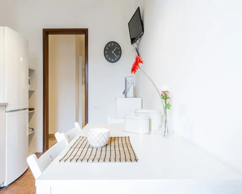 Angolo cucina luminoso e minimale con piano da pranzo bianco, tovaglietta decorativa e vaso con fiori—stile moderno e pulito, ideale per piccoli appartamenti.