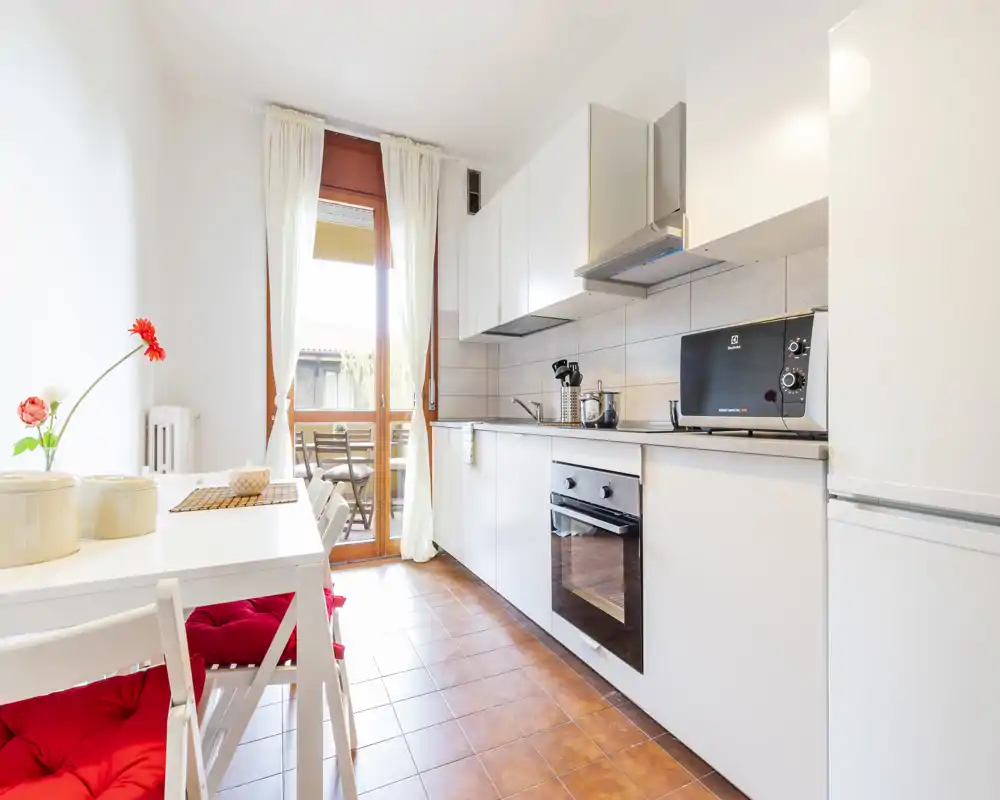 Cucina moderna e luminosa con mobili bianchi, forno integrato e un piccolo tavolo da pranzo vicino alla porta del balcone che illumina l'ambiente.