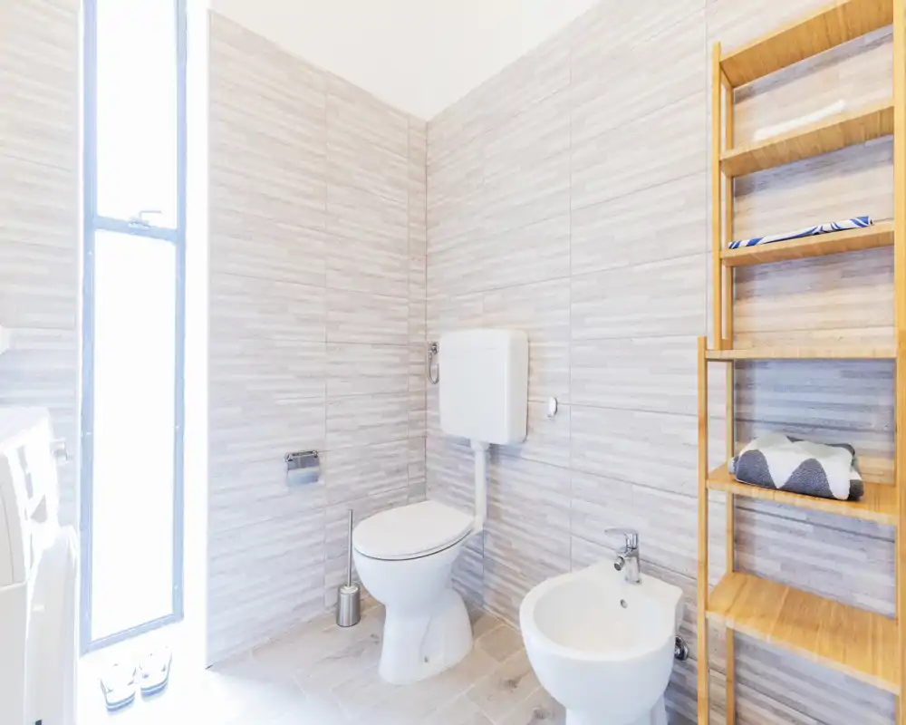Bagno luminoso e moderno con WC, bidet e scaffalatura in legno; pareti piastrellate e una finestra alta satinata per luce naturale.