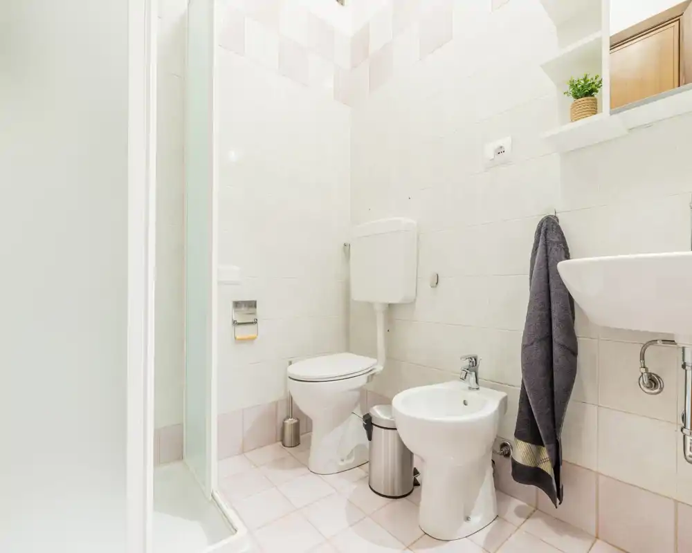 Bagno luminoso e ben tenuto con box doccia, WC, bidet e lavabo a parete. Piastrelle neutre e accessori ordinati danno una sensazione pulita e funzionale.
