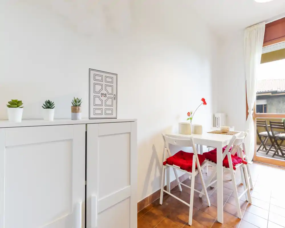 Accogliente zona pranzo luminosa con un piccolo tavolo bianco e quattro sedie con cuscini rossi, luce naturale da un balcone e decorazioni minimal.
