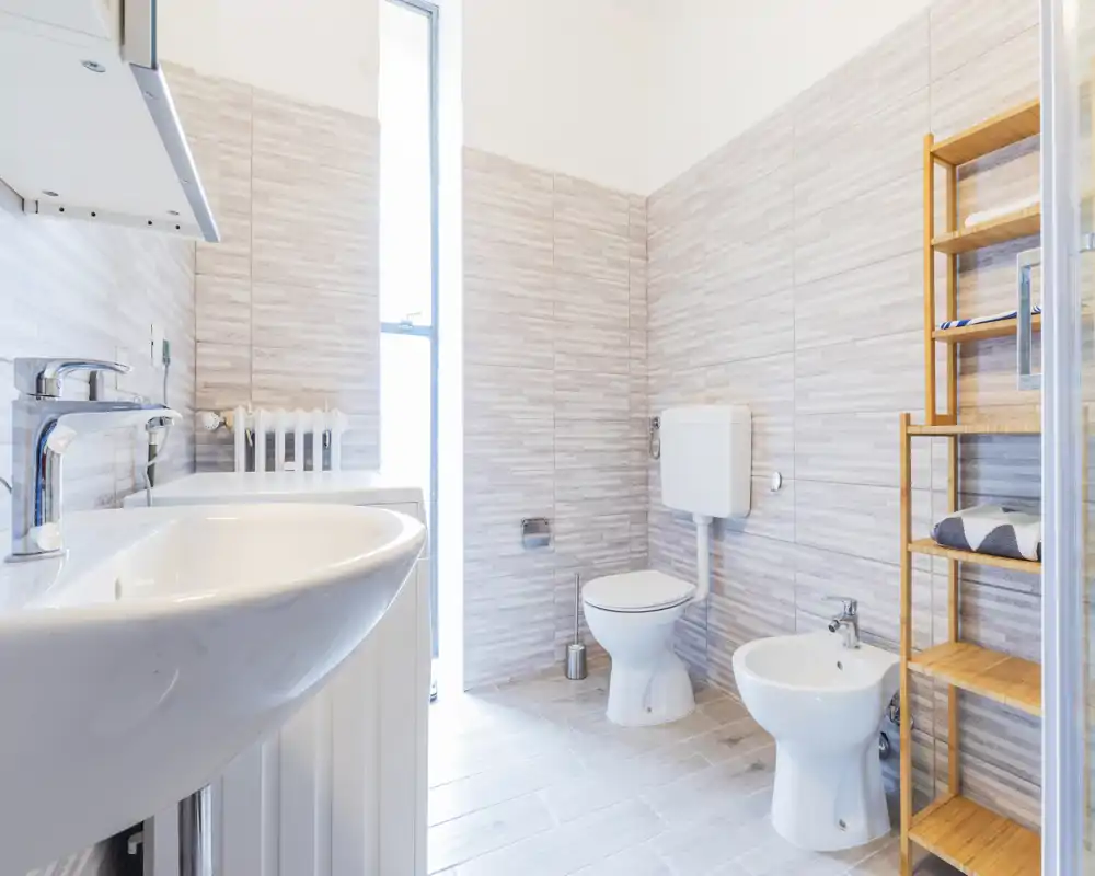 Bagno moderno e luminoso con lavabo a parete, wc e bidet, piastrelle chiare e un mobiletto in legno per asciugamani e riporre oggetti.
